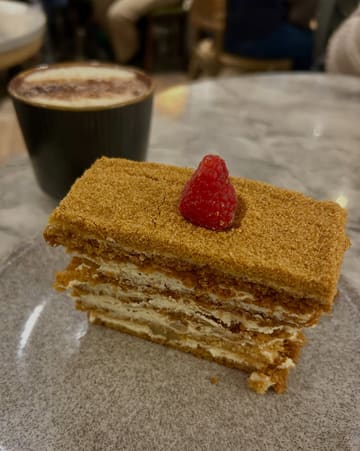 A close-up, eye-level shot of a slice of honey cake served on a grey plate, with a Hojicha latte visible in the background. The image captures a casual cafe setting, focusing on the layered texture of the cake.