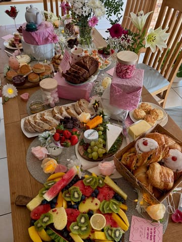 A high-angle, overhead shot of a lavish grazing table spread, featuring a variety of pastries, scones, cheeses, and fresh fruit, decorated with flowers and candles for a hen party celebration.