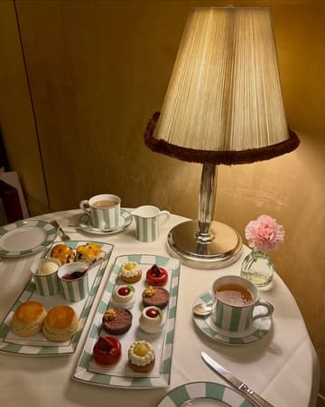 A beautifully composed, atmospheric photograph of an afternoon tea service at Claridge's. The image captures a selection of delicate pastries, scones with accompaniments, and tea, arranged elegantly on a table with warm, ambient lighting that highlights the luxury setting.