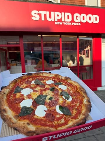 A casual, first-person perspective shot of a pizza box held in front of a restaurant storefront. The image features a pepperoni and burrata pizza, showcasing the crust and toppings, with the venue's branding visible in the background.