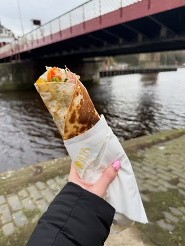 A first-person perspective shot of a lamb kofta wrap held in front of a river, capturing a casual street food experience at a market.