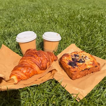 A casual, sunny-day picnic shot featuring a croissant and a blueberry pastry on paper bags placed directly on the grass, accompanied by two takeaway coffee cups.