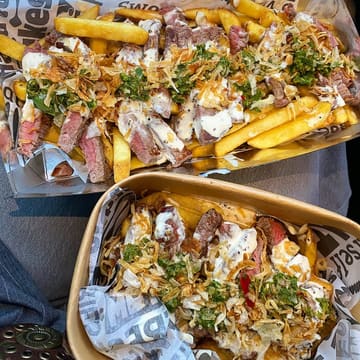 A top-down shot of two takeaway containers filled with steak and chips, presented on a lap inside a car. The lighting is bright and natural, highlighting the textures of the seared meat, crispy chips, and toppings.