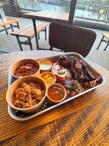 A high-angle shot of a large BBQ platter served on a metal tray, featuring various meats, beans, and sides, set on a wooden table with a view of the waterfront in the background.