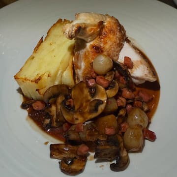 A close-up, top-down shot of a Pan-Fried Chicken Supreme dish served with Dauphinoise potatoes and a coq au vin style sauce. The image captures the texture of the chicken skin and the mushrooms, though the lighting is harsh.
