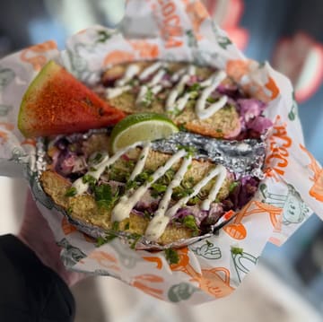 A close-up, top-down shot of a Mexican torta served in a branded paper-lined basket, held by a person. The image captures the messy, casual nature of the street food, highlighting the ingredients like red cabbage, cilantro, and lime.