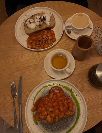 A top-down, dimly lit photograph of a brunch meal consisting of beans on sourdough toast with a green herb oil drizzle, accompanied by a coffee and a cup of tea, captured in a cafe setting.