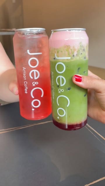A simple, casual shot of two hands holding branded iced drinks from a cafe, featuring a strawberry iced tea and a strawberry matcha latte. The composition is straightforward, focusing on the product branding, though the framing is slightly tight.