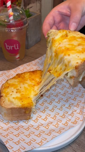 A close-up, first-person perspective shot of a grilled cheese sandwich being pulled apart to show the melted cheese, with a branded iced drink visible in the blurred background.
