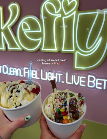 A first-person perspective shot holding two cups of frozen yogurt with various toppings in front of a neon-lit brand sign.