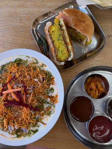 A top-down shot of a casual Indian meal featuring samosa chaat, a vada pav, and a selection of chutneys and pickles served on a wooden table.