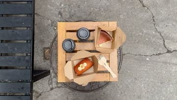 A top-down, flat-lay shot of takeaway coffee and pastries placed on a wooden crate, which is situated on a manhole cover.