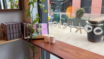A simple, atmospheric shot of two takeaway coffee cups sitting on a wooden window ledge in a cafe, with a view of the street outside.