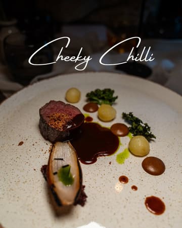 A moody, close-up shot of a fine-dining plated dish, likely a piece of beef with jus and garnishes, presented in a dark, atmospheric setting.