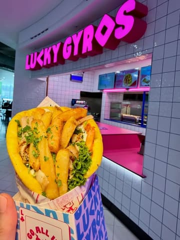 A close-up, handheld shot of a gyro held in front of the Lucky Gyros stall in the Manchester Arndale food court, showcasing the branding and the product.