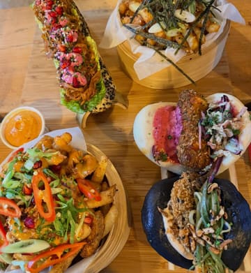 A top-down flat lay shot of a spread of bao buns and loaded fries served on a wooden board. The image captures a variety of dishes including different types of bao buns and loaded fries, presented in bamboo steamers and on a wooden surface.