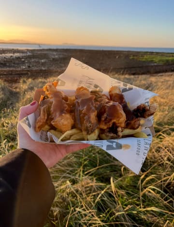 A handheld, close-up shot of deep-fried pigs in blankets topped with gravy, served in branded paper, captured against a scenic Scottish beach at sunset.