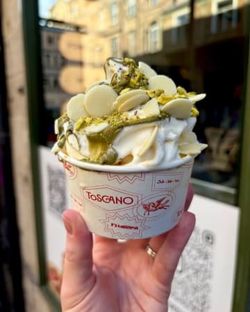 A close-up, handheld shot of a pistachio soft-serve gelato topped with white chocolate buttons, served in a branded paper cup. The image is taken at a 45-degree angle, effectively showcasing the texture and toppings of the dessert against a softly blurred street background.