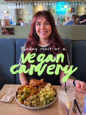 A medium shot of a woman sitting at a restaurant table with a large plate of a vegan Sunday roast in front of her, featuring a prominent green text overlay.