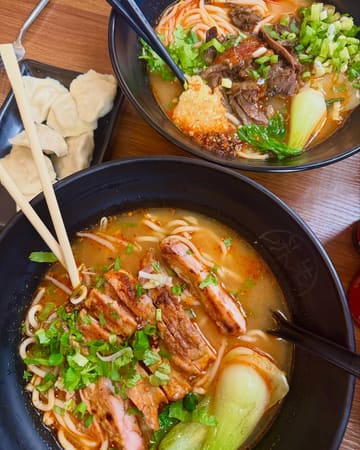 A top-down, overhead shot of two bowls of noodle soup and a side of dumplings served on a wooden table. The image focuses on the textures of the meat and the broth, presenting a casual dining experience.
