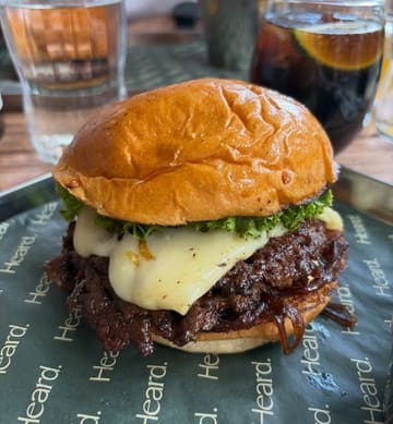 A close-up, eye-level shot of a smash burger served on branded paper, featuring a melted cheese layer and caramelized onions. The image captures the texture of the patty and the bun, though the composition is somewhat tight.