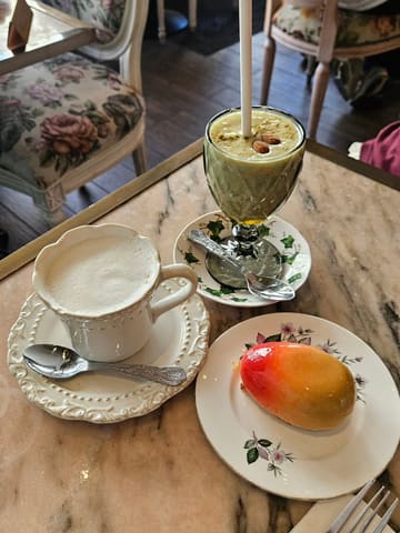 A top-down shot of a table at a cafe featuring a mango-shaped mousse cake, a green avocado-based drink, and a cup of coffee, served on floral china.