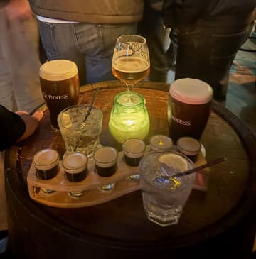 A casual, atmospheric shot taken at an Irish bar, featuring a selection of drinks including Guinness and baby Guinness shots arranged on a barrel table. The lighting is dim and moody, typical of a pub environment, with a candle providing a warm focal point.