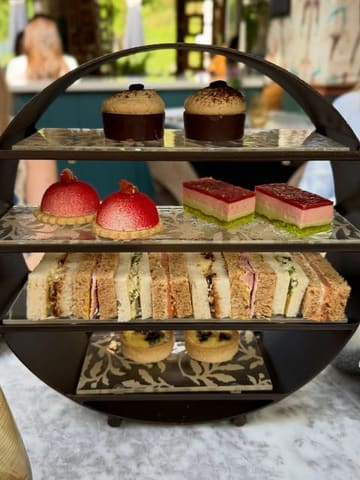 A high-quality, well-composed shot of an afternoon tea stand at a premium hotel. The lighting is soft and natural, highlighting the textures of the pastries and sandwiches, while the shallow depth of field keeps the focus entirely on the food.