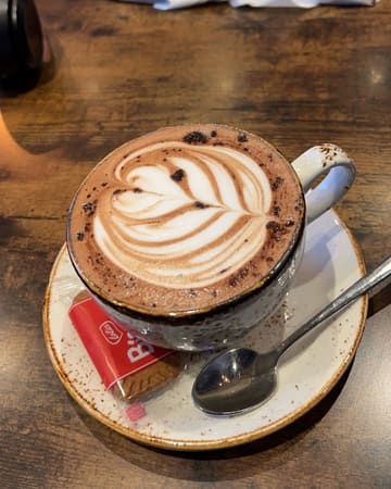 A clear, well-composed close-up shot of a cappuccino featuring latte art, served on a saucer with a Lotus Biscoff biscuit on a dark wooden table.