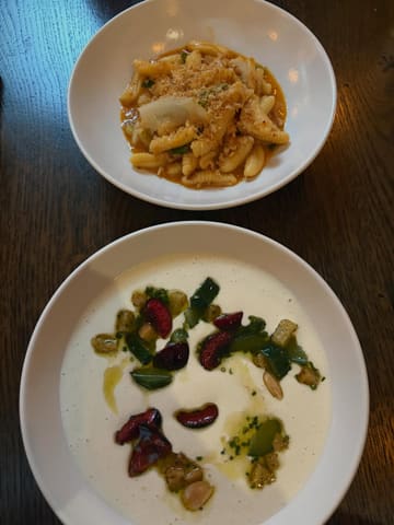 A top-down, overhead shot of two distinct dishes served at a restaurant, featuring a pasta dish and a white soup with dark garnishes, set against a dark wooden table.