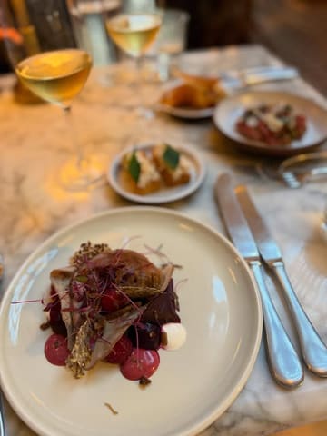 A high-quality, atmospheric shot of a fine dining spread at The Spence in Edinburgh, featuring a beautifully plated main dish in the foreground with drinks and other courses softly blurred in the background.