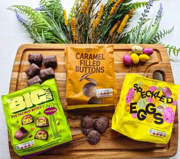 A clean, well-lit flat-lay photograph showcasing M&S chocolate products, including 'The Big Daddy' Pistachio Bites, Caramel Filled Buttons, and Speckled Eggs, arranged on a wooden board with decorative dried flowers.