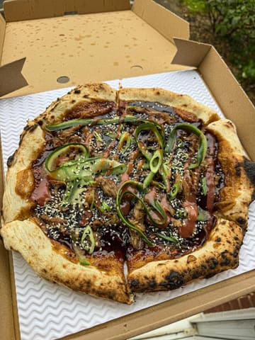 A top-down, eye-level shot of a Hoisin Duck pizza served in a cardboard takeaway box. The pizza features a wood-fired crust, dark sauce, cucumber ribbons, spring onions, and sesame seeds.