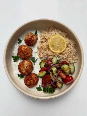 A clean, top-down, studio-style photograph of a homemade meal featuring Greek-style turkey meatballs, a generous dollop of tzatziki, a fresh tomato and cucumber salad, and a portion of rice. The lighting is bright and even, highlighting the textures of the food, and the presentation is simple and appetizing.