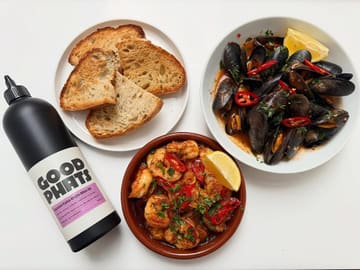 A high-quality, well-lit flat lay photograph featuring a bottle of 'Good Phats' Spanish extra virgin olive oil alongside a meal of gambas pil pil, harissa mussels, and crusty sourdough bread. The composition is clean and professional, effectively showcasing the product in a culinary context.