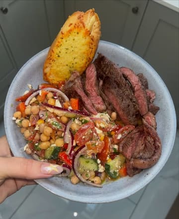A top-down shot of a home-cooked meal prep bowl containing a chickpea and mixed bean salad, topped with sliced steak and a piece of garlic bread.