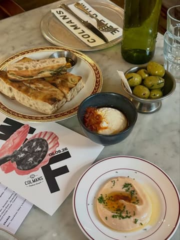 A high-quality, overhead flat-lay photograph capturing a diverse spread of dishes from a collaborative dining event. The composition is well-balanced, showcasing various small plates, dips, and bread, with the event menu clearly visible to provide context. The lighting is soft and natural, highlighting the textures of the food, and the focus is sharp across the entire table.