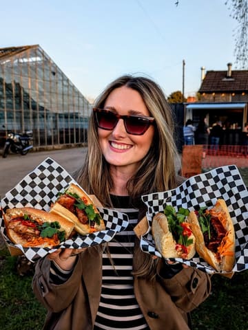 A bright, high-quality promotional shot featuring a smiling woman holding two vibrant Bánh Mì sandwiches in a casual, outdoor brewery setting. The image effectively captures the street food vibe and the relaxed atmosphere of the venue.