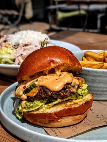 A high-quality, close-up food photography shot of a double smash burger with melted cheese and sauce, served with fries and a salad in the background. The image captures a casual, appetizing lunch setting, likely outdoors.