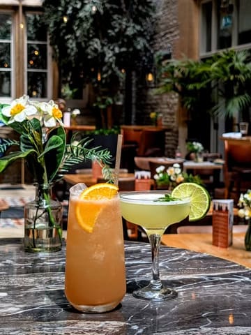 A high-quality, well-lit photograph of two cocktails on a marble table, set within the bright, airy, and slightly cluttered interior of a hotel conservatory or restaurant.