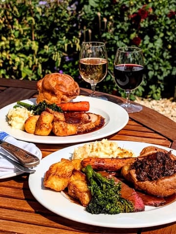 A bright, outdoor shot of two Sunday roast dinners served on a wooden table, likely in a pub garden. The image captures the classic British pub aesthetic with roast meat, Yorkshire puddings, and seasonal vegetables, accompanied by glasses of wine.