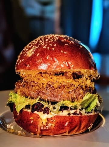 A high-quality, close-up, eye-level shot of a gourmet burger, showcasing distinct layers of ingredients including a patty, melted cheese, lettuce, and black pudding, served on a toasted brioche bun.