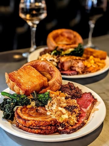 A high-quality, appetizing shot of a Sunday roast at Bath Chophouse, featuring a well-composed foreground plate with sirloin, porchetta, and beef shin, with a second plate blurred in the background to create depth.