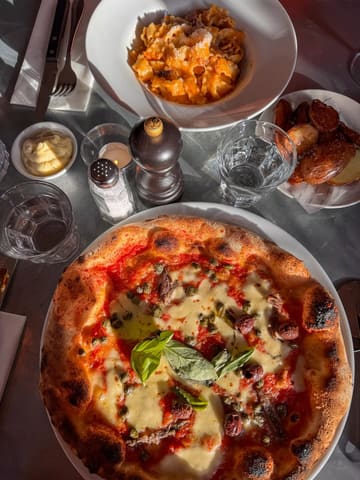 A high-quality, overhead flat-lay shot of a restaurant table featuring a Neapolitan-style pizza, a bowl of pasta, and a side of roasted potatoes. The natural sunlight creates a warm, inviting atmosphere, highlighting the textures of the food.
