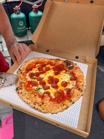 A high-angle shot of a freshly baked Neapolitan-style pepperoni pizza in a cardboard box, being sliced by a pizza cutter. The setting appears to be a casual outdoor pop-up environment, evidenced by the gas canisters in the background.