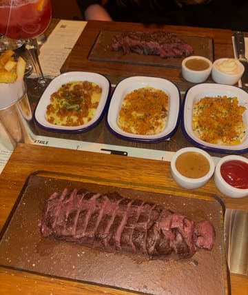A top-down, eye-level shot of a meal at Flat Iron, featuring a sliced flat iron steak on a wooden board, accompanied by various sides including mac and cheese, mash, and fries, along with sauces and a cocktail.