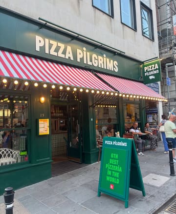 A clear, daylight photograph of the Pizza Pilgrims storefront on Corn Street, featuring the restaurant's signage, awning, and outdoor seating area.