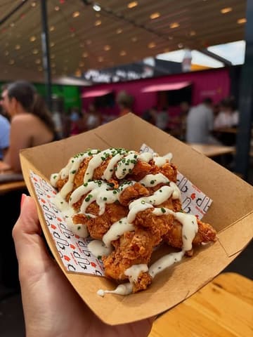 A close-up, first-person perspective shot of a cardboard tray filled with crispy fried chicken strips drizzled with a creamy garlic herb sauce, held in a hand at an outdoor street food venue.