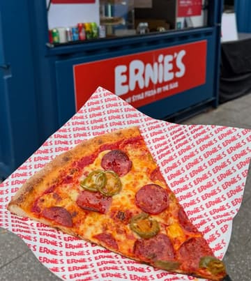 A close-up, handheld shot of a single slice of pepperoni and jalapeño pizza, held against the backdrop of the 'Ernie's' street food container.