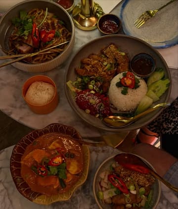 A top-down, dimly lit shot of a table spread featuring various Thai dishes including udon noodles, panang curry, sticky rice, and roasted meats. The image captures a casual dining experience with a warm, moody atmosphere typical of a restaurant setting.